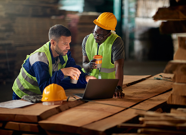 Two carpentry contractors having coffee while looking at blueprints on a laptop