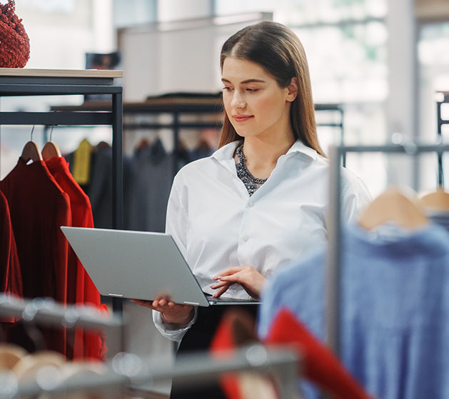 A clothing store attendant standing by clothing racks looking over her online store's website secruity on her iPad